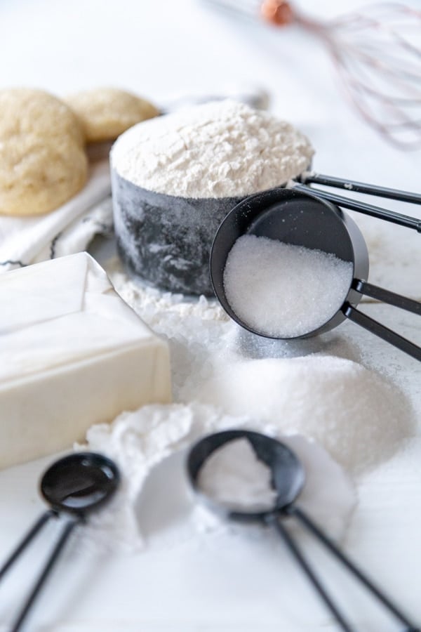 The ingredients for sugar cookies in black measuring cups and spoons on a white wood board. 