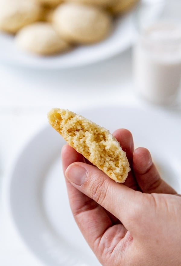 A hand holding half of a sugar cookie over a white plate with a platter of cookies and a glass of milk.