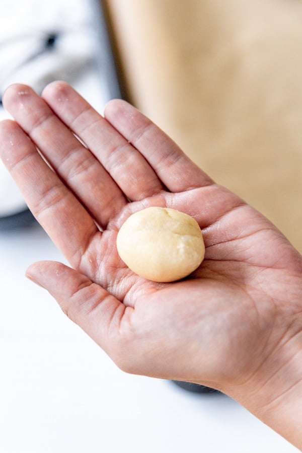 A hand holding a ball of cookie dough over a baking sheet.