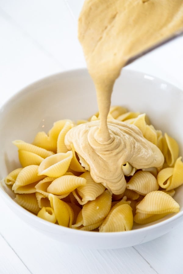 Cheese sauce being poured over pasta shells in a white bowl. 