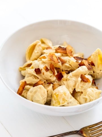 A white bowl with mac and cheese and coconut bacon sprinkled on top with a silver fork next to the bowl.