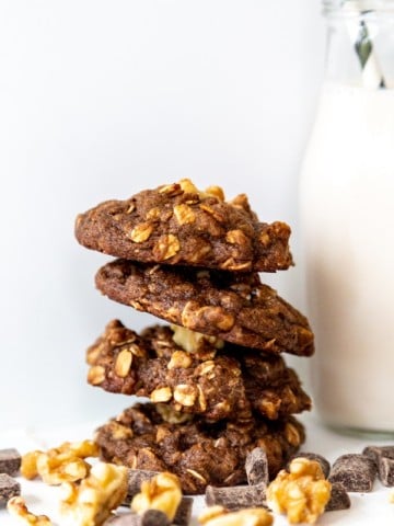 Four double chocolate chip cookies stacked with walnuts and a glass of milk with a straw in it