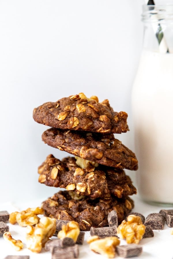 Four double chocolate chip cookies stacked with walnuts and a glass of milk with a straw in it