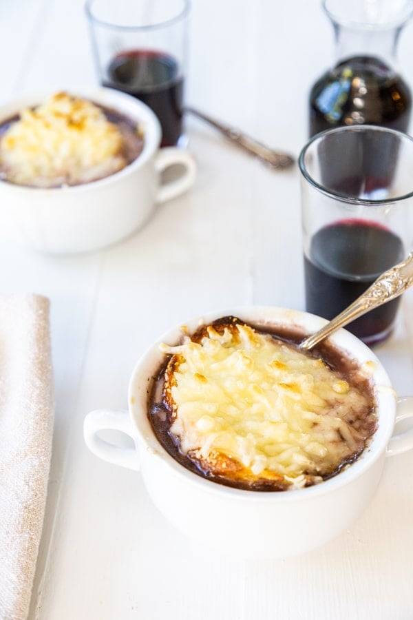 Two bowls of French onion soup with glasses of red wine and spoons in the bowls.