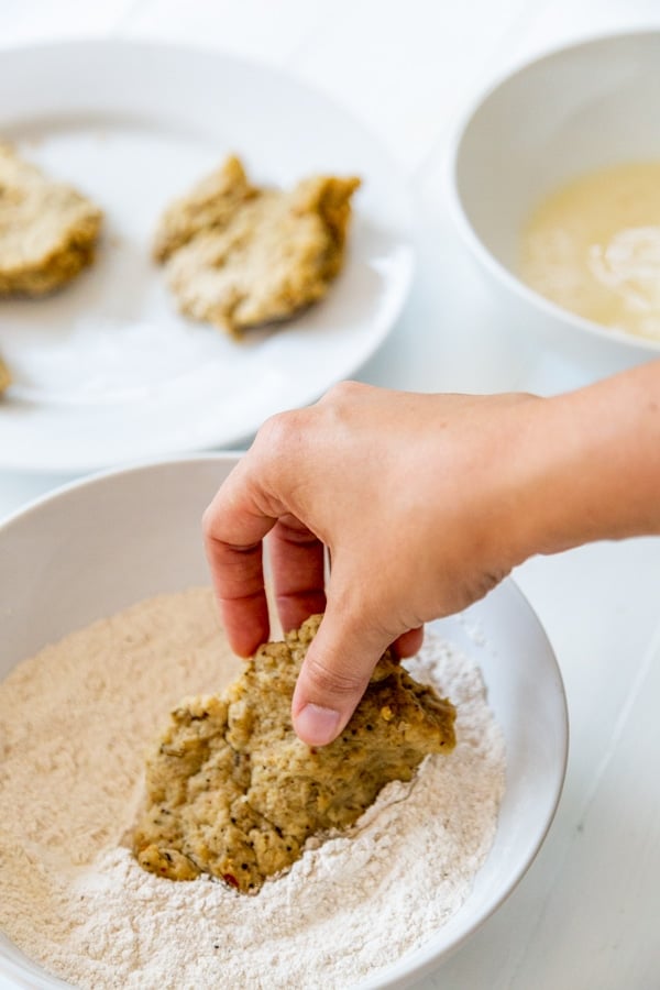 A hand dipping a chicken cutlet in a bowl of flour.