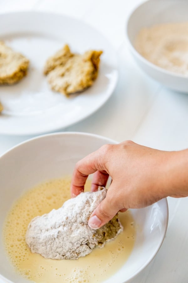 A hand dipping a floured chicken cutlet in a bowl of eggs.