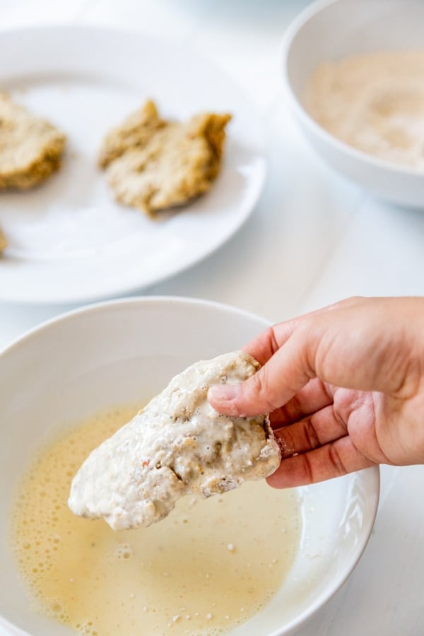 A hand dipping a chicken cutlet coated in flour and egg in a bowl of flour.