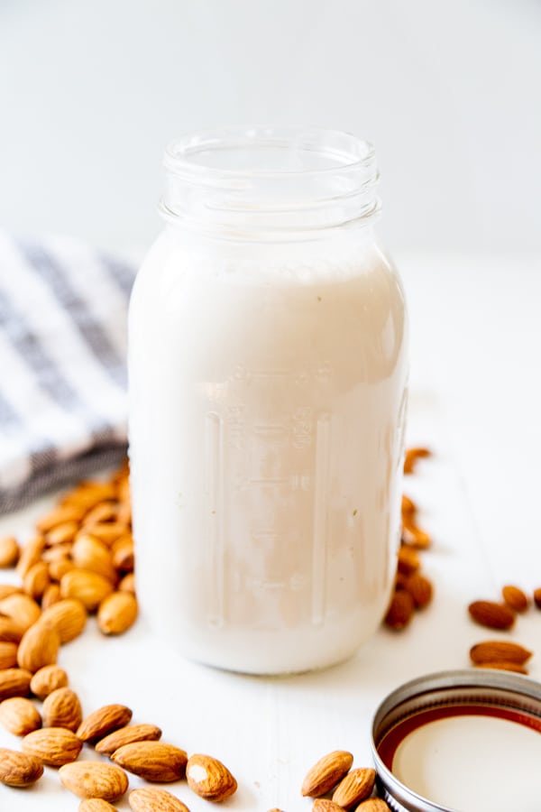 A jar of almond milk with almonds scattered next to it on a white wood table.