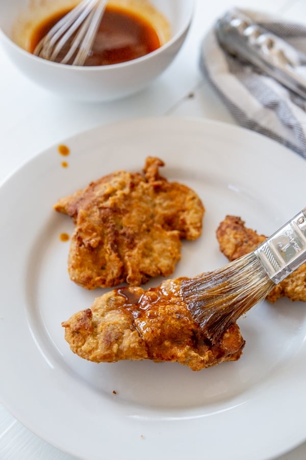 A pastry brush putting hot chicken sauce on fried chicken on a white plate.