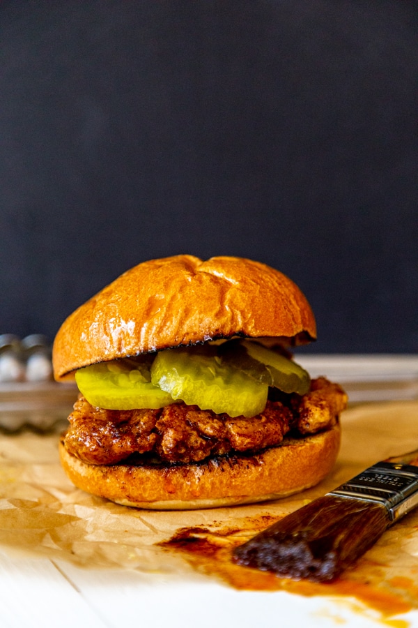 A spicy fried chicken sandwich with pickles on parchment paper with a black background. 