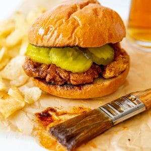 A spicy fried chicken sandwich on parchment paper with a pastry brush dripping spicy sauce next to it.