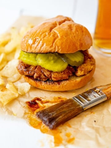 A spicy fried chicken sandwich on parchment paper with a pastry brush dripping spicy sauce next to it.