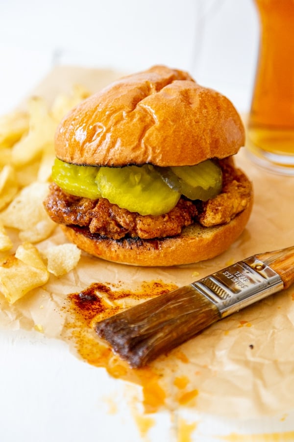 A spicy fried chicken sandwich on parchment paper with a pastry brush dripping spicy sauce next to it. 