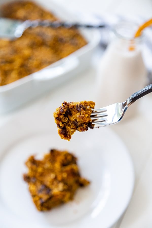 A blondie on a white plate with the pan of blondies in the background and a fork with a piece of blondie.