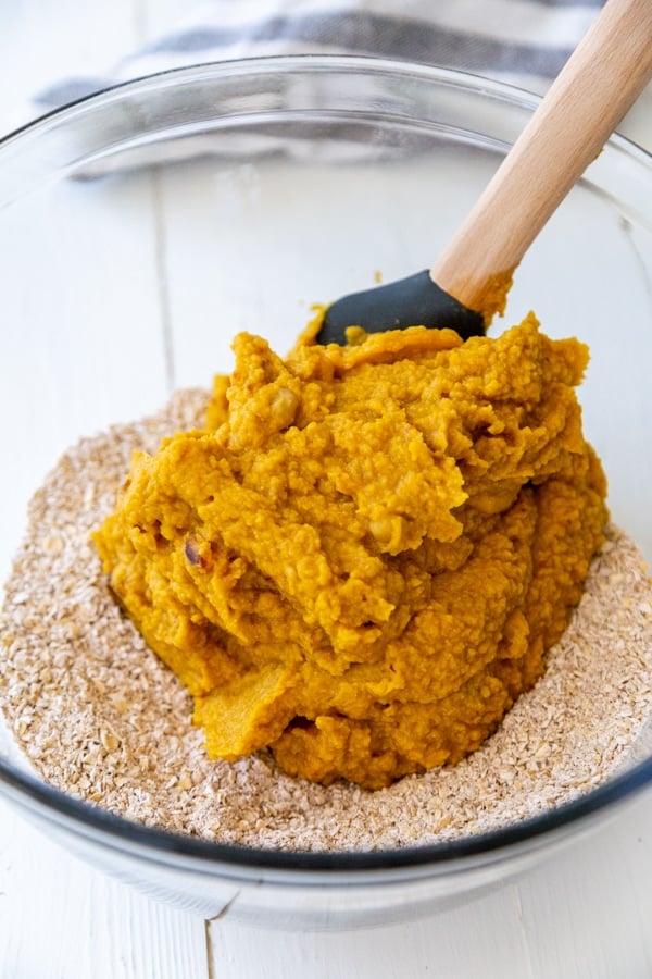 A glass mixing bowl with oat flour and a spatula with pumpkin puree.