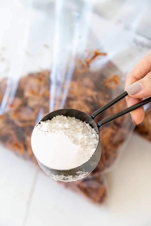 A hand holding a black measuring cup of powdered sugar over a bag of chocolate cereal.