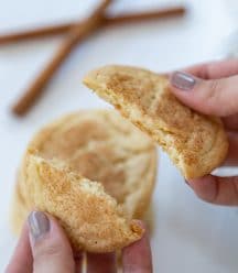 A hand breaking a cookie in half over a stack of cookies and cinnamon sticks.