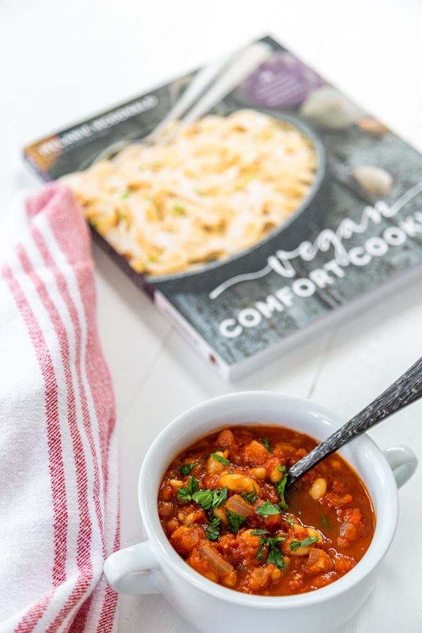 A white bowl with tomato soup and a silver spoon and a cookbook and towel in the background. 