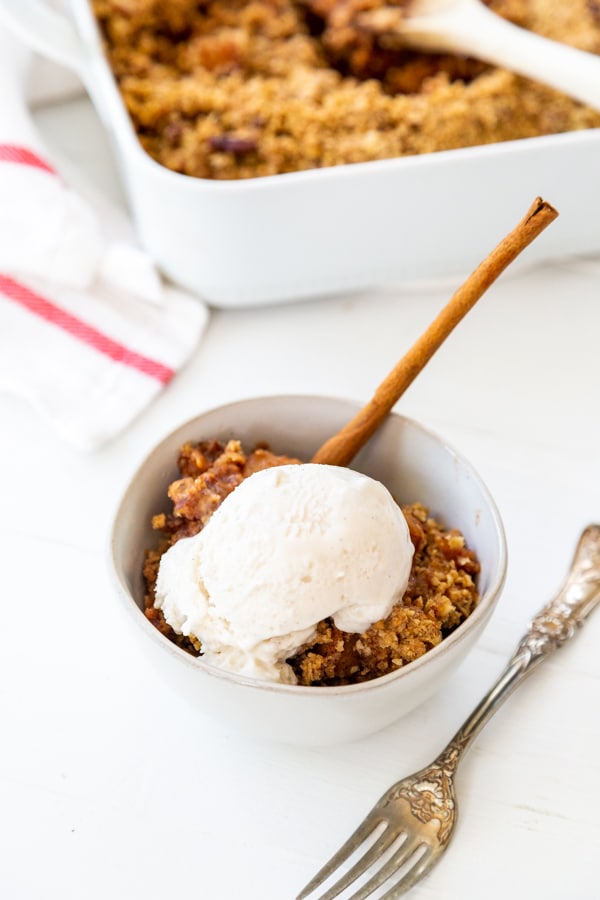 A bowl of apple crisp with ice cream on top and a pan of crisp behind it. 