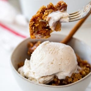 A bowl of apple crisp with ice cream and a fork taking a bite of the crisp out of the bowl.