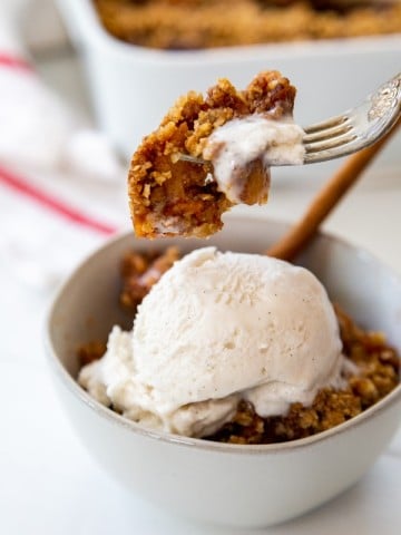 A bowl of apple crisp with ice cream and a fork taking a bite of the crisp out of the bowl.