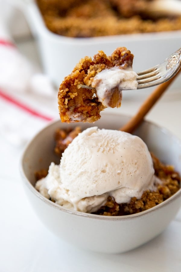 A bowl of apple crisp with ice cream and a fork taking a bite of the crisp out of the bowl.
