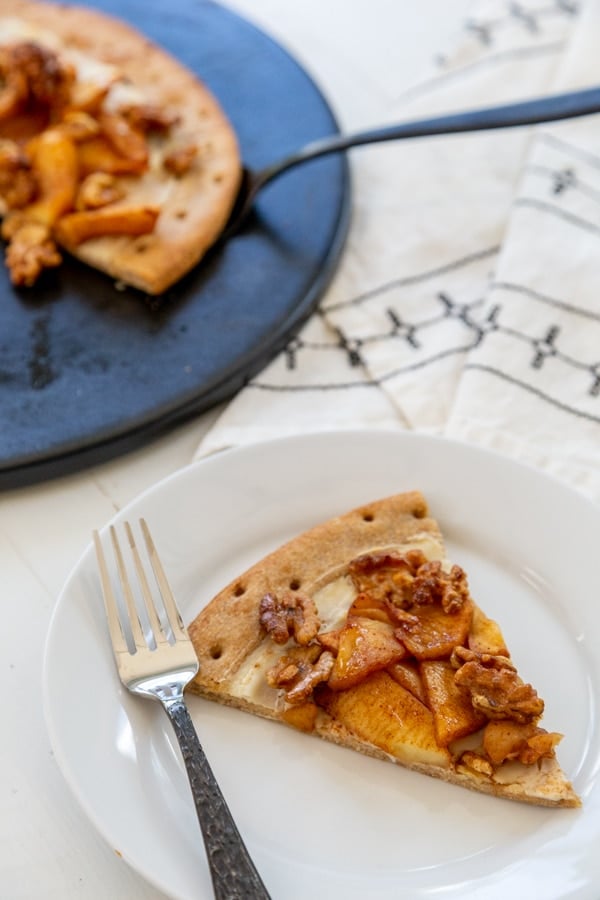 A slice of apple pie pizza on a white plate with the rest of the pizza behind it on a black pizza stone.