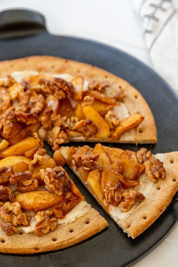 An apple pie pizza with a slice cut out of the pie on a black pizza stone. 