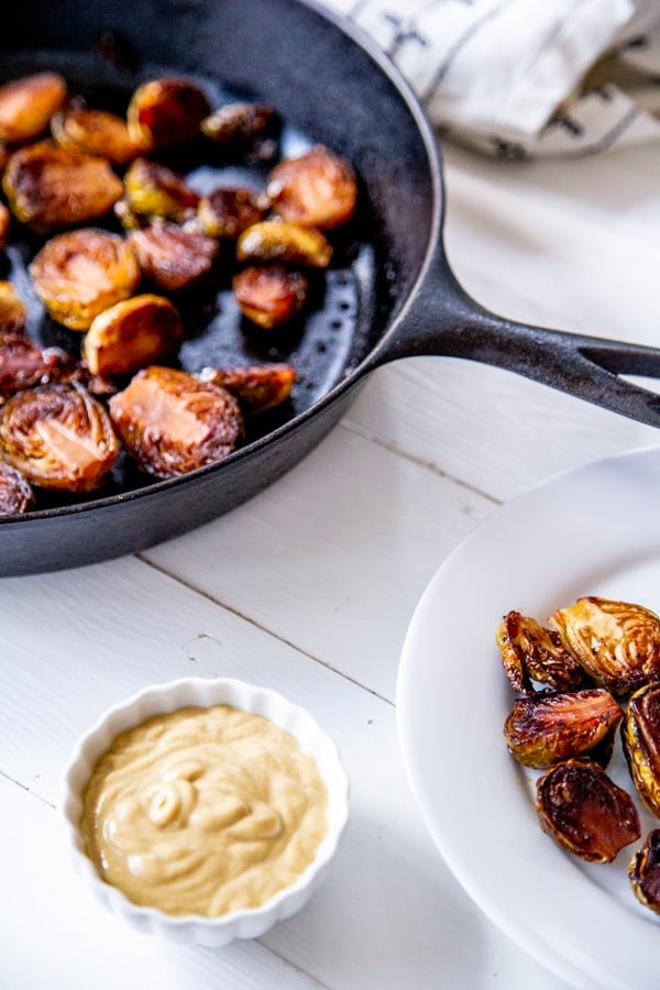 An iron skillet with roasted Brussels sprouts and a white plate of sprouts and a white bowl of dip.