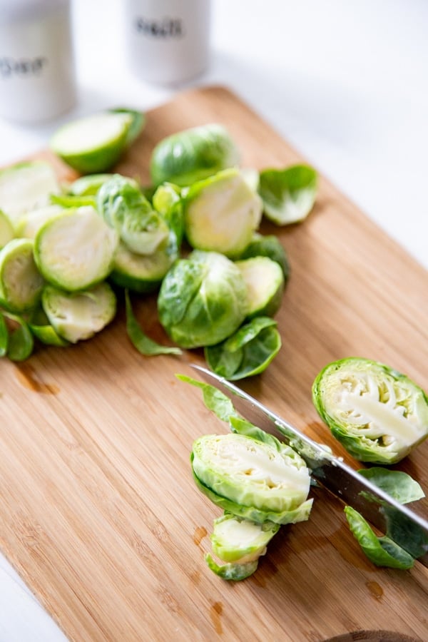 A knife with a Brussels sprout cut in half on a wood board with more halved sprouts behind it.