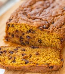 A loaf of pumpkin bread with chocolate chips and a slice off of the end on a wood board.