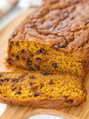 A loaf of pumpkin bread with chocolate chips and a slice off of the end on a wood board.