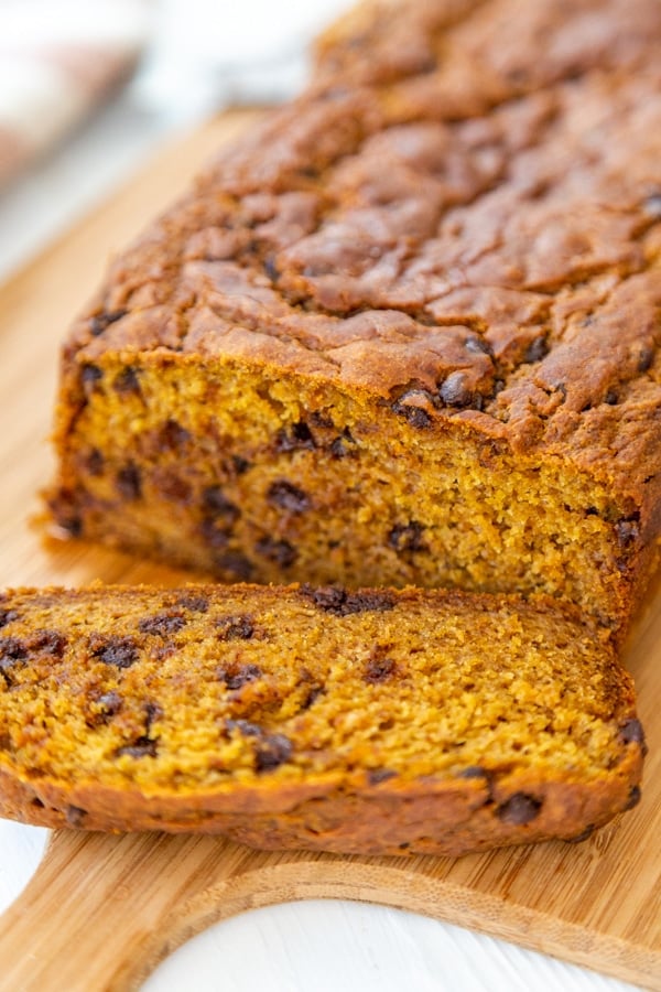 A loaf of pumpkin bread with chocolate chips and a slice off of the end on a wood board.