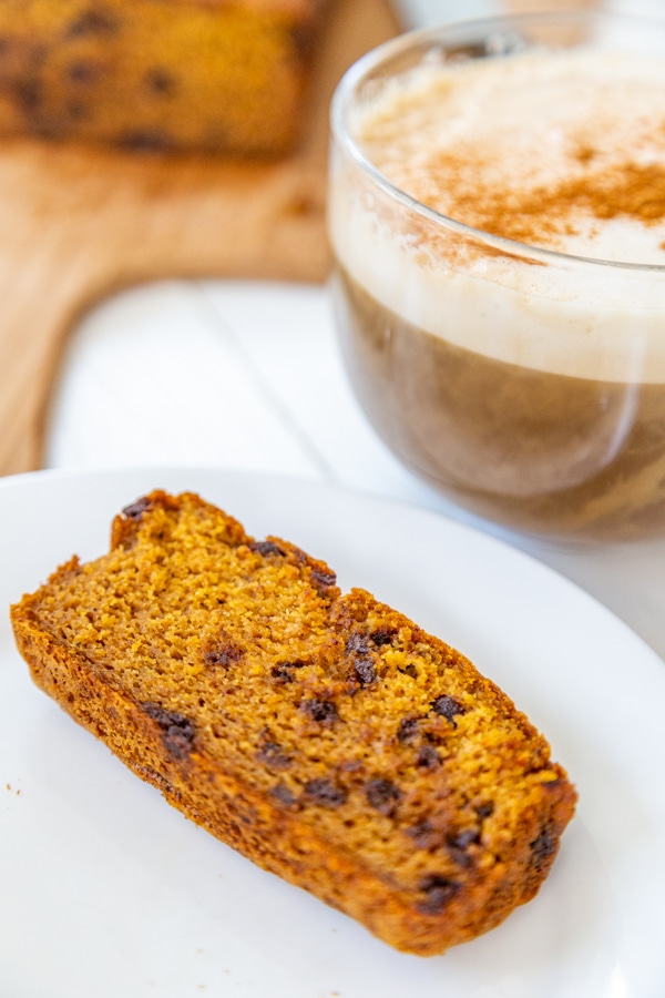 A slice of chocolate chip pumpkin bread on a white plate with a cup of coffee next to it. 