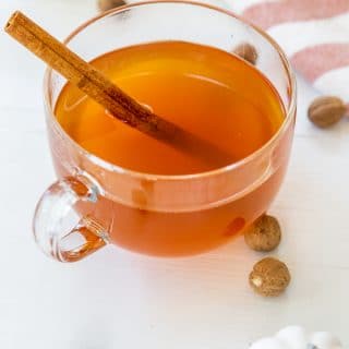 A clear glass mug of mulled apple cider with a cinnamon stick and white pumpkins, spices, and a towel on a white table.
