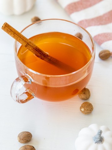 A clear glass mug of mulled apple cider with a cinnamon stick and white pumpkins, spices, and a towel on a white table.