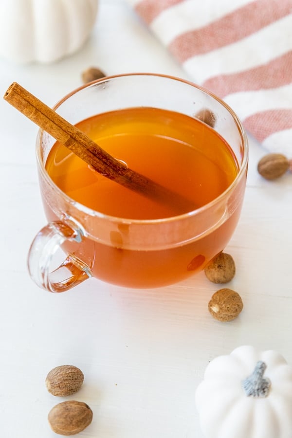 A clear glass mug of mulled apple cider with a cinnamon stick and white pumpkins, spices, and a towel on a white table. 