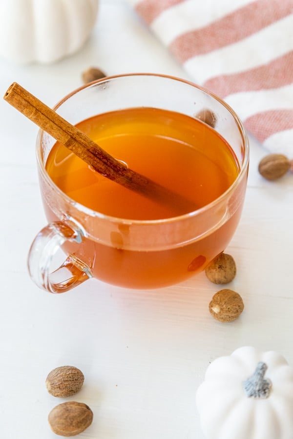 A clear glass mug of mulled apple cider with a cinnamon stick and white pumpkins, spices, and a towel on a white table.