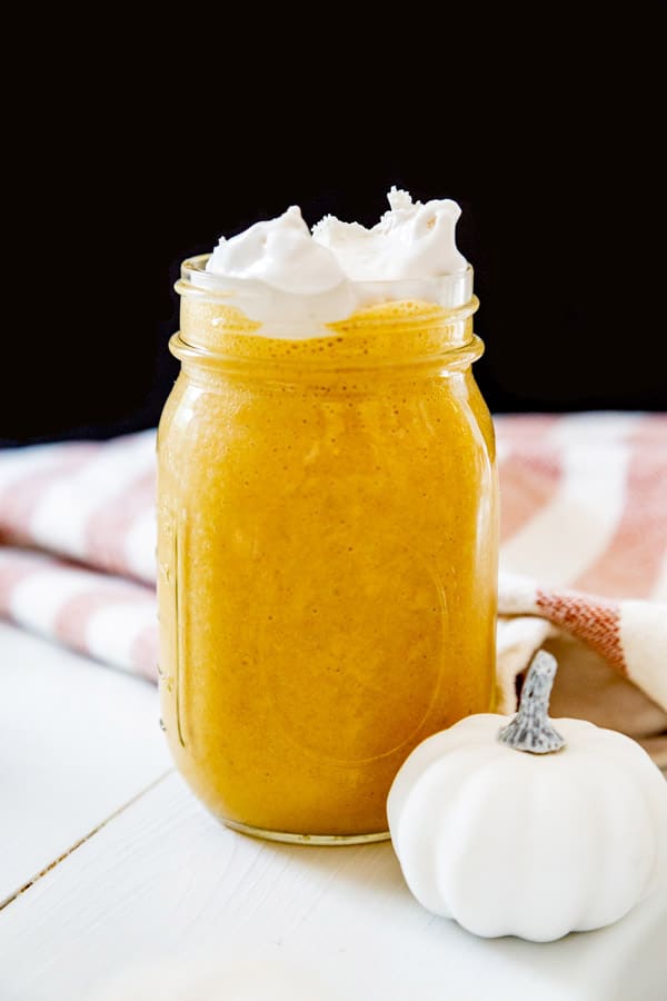A pumpkin smoothie with whipped cream in a mason jar and a white pumpkin next to the jar. 