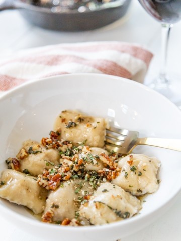 A white bowl of ravioli with brown butter and pecans on top.