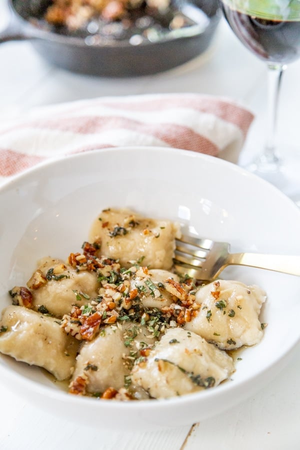 A white bowl of ravioli with brown butter and pecans on top. 