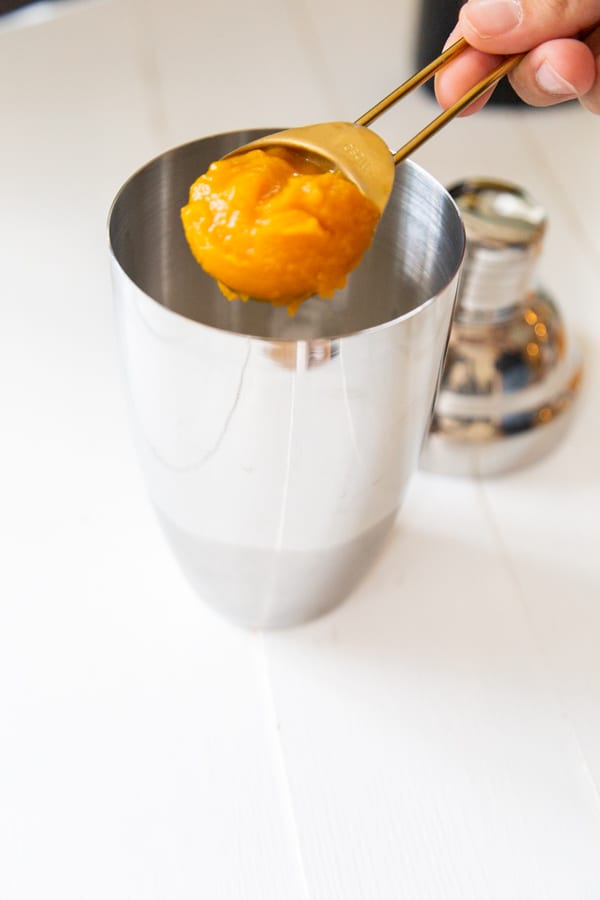 A hand putting a spoonful of pumpkin puree into a martini shaker.