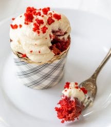 A red velvet cupcake with cream cheese frosting on a white plate with a fork full of the cake resting on the plate.