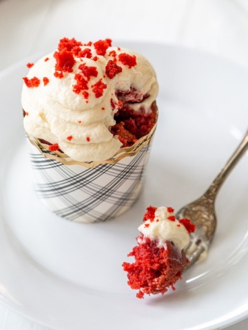 A red velvet cupcake with cream cheese frosting on a white plate with a fork full of the cake resting on the plate.