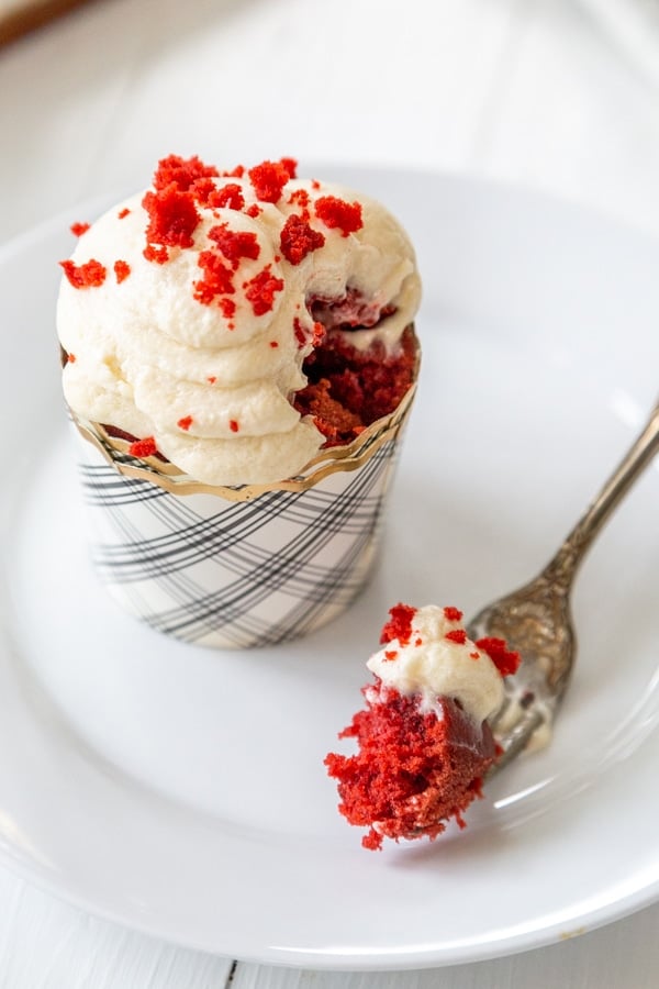 A red velvet cupcake with cream cheese frosting on a white plate with a fork full of the cake resting on the plate.