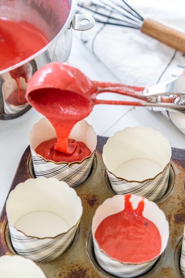 A hand filling a cupcake liner with red velvet cake batter.