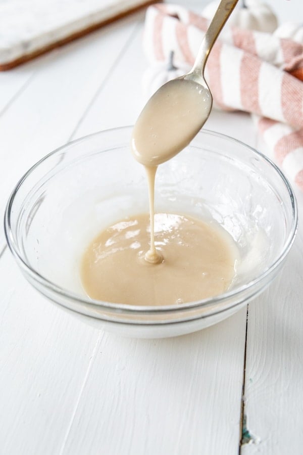 A glass bowl with icing and a spoon dripping icing in the bowl.