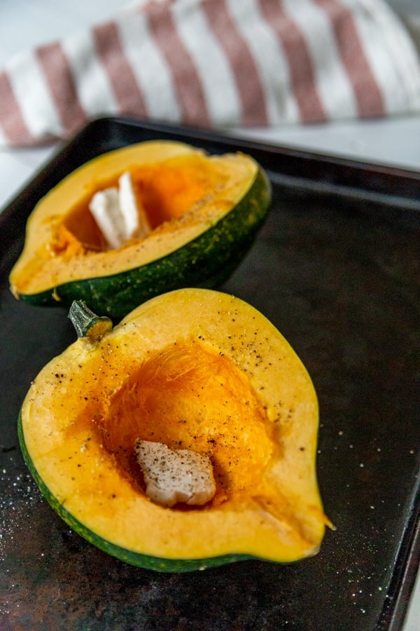 Two halves of an acorn squash on a baking pan with butter, salt, and pepper.