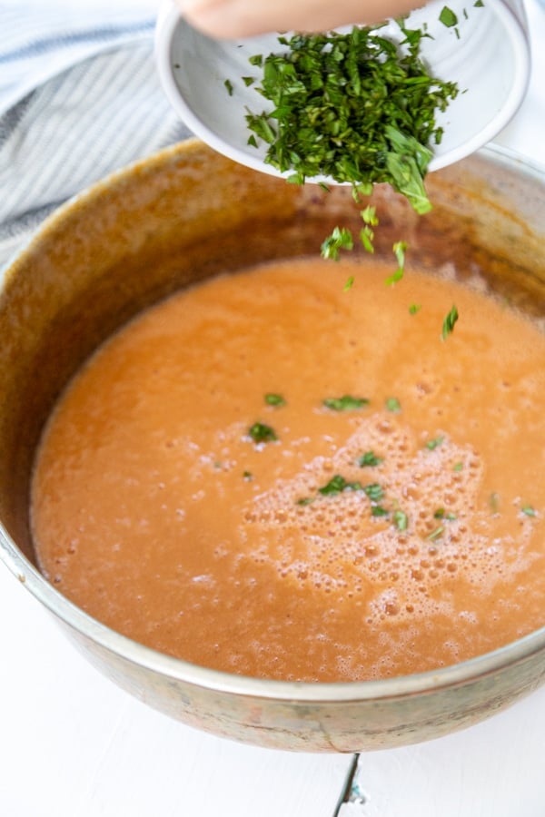 A copper pot of tomato soup with minced basil being sprinkled on top. 