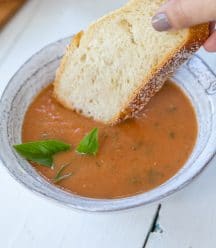 A bowl of tomato soup with a hand dipping a slice of bread in it.